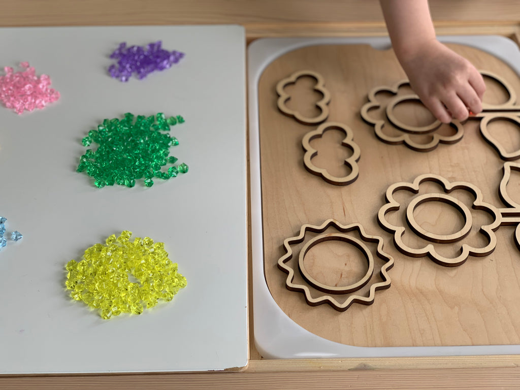 Fillable flower-themed wooden FLISAT table insert with child placing petal pieces and colorful translucent gems nearby