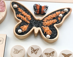 Natural wooden butterfly-shaped sensory tray filled with orange and black rice and white felt balls on white background