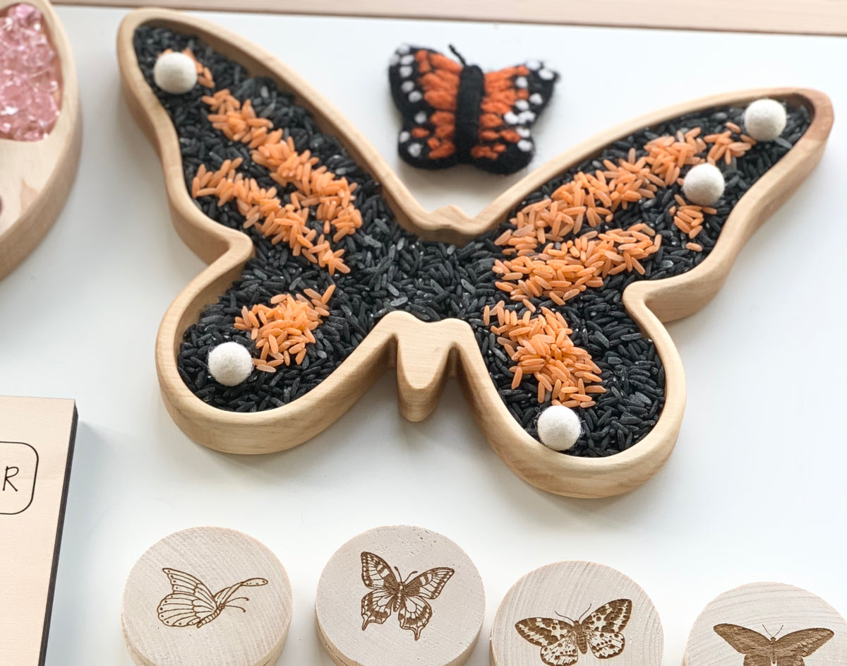 Natural wooden butterfly-shaped sensory tray filled with orange and black rice and white felt balls on white background