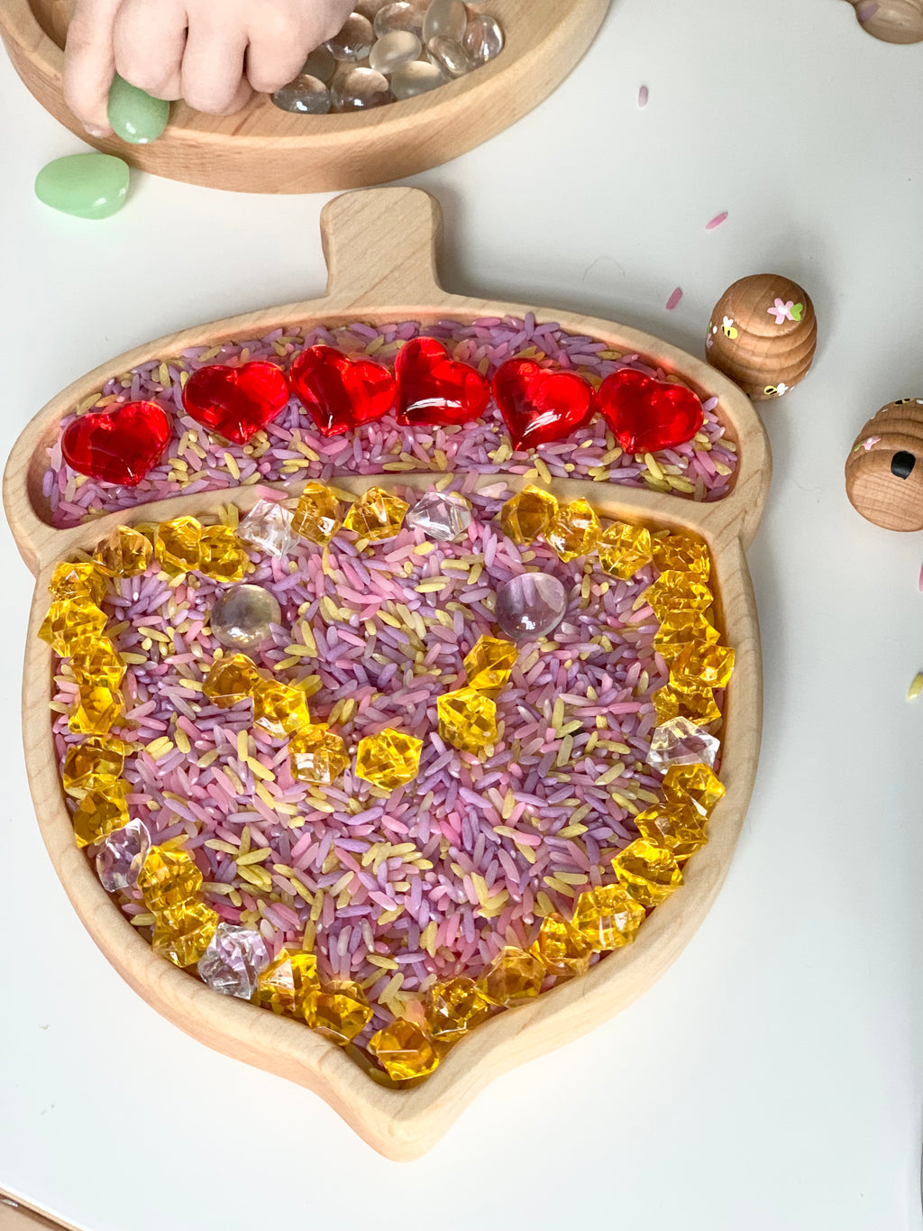 Acorn-shaped wooden sensory tray filled with colored rice, red heart gems, and yellow crystal-like beads