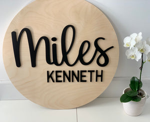 3D round wooden name sign with black cursive and block letters on natural wood background near white orchid plant