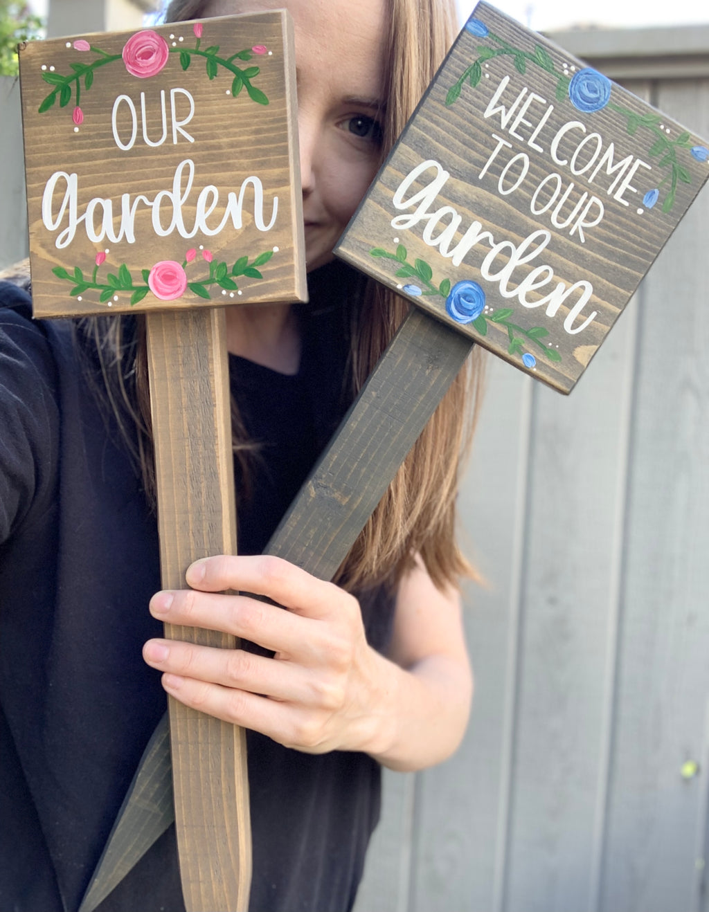 Two small square wooden garden signs with floral accents held by a person against a fence background