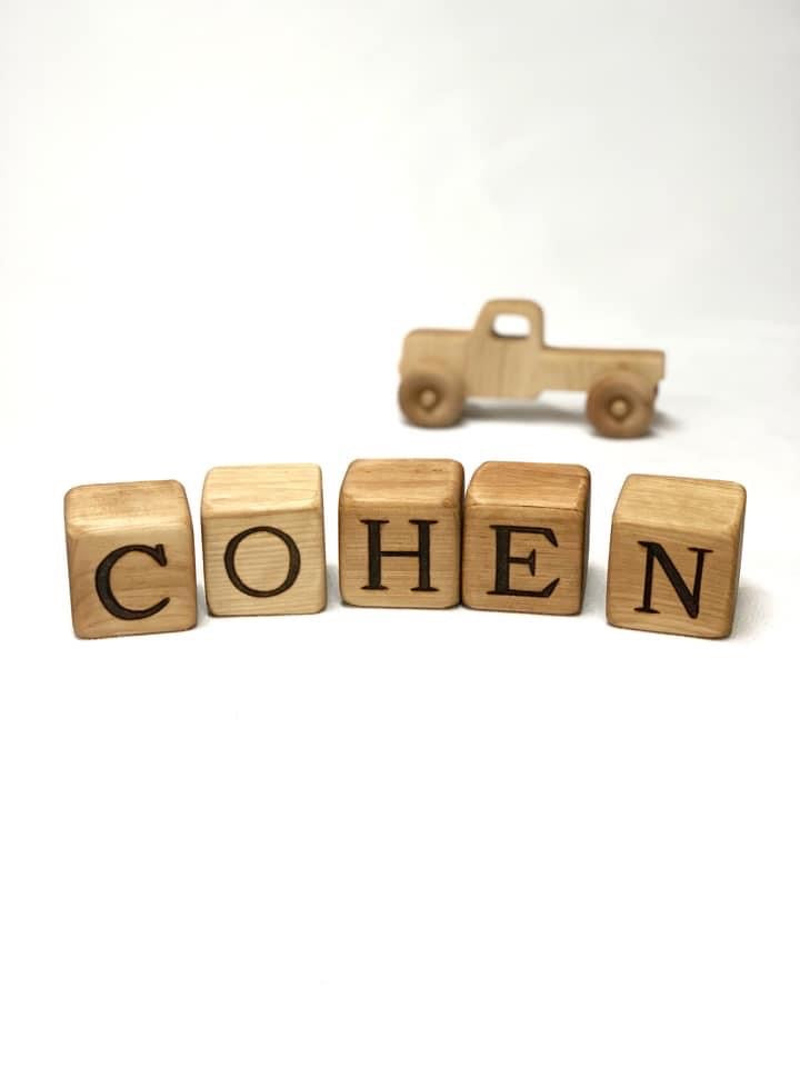 Custom 2 inch wooden blocks spelling COHEN with a small wooden toy truck in the background on white surface