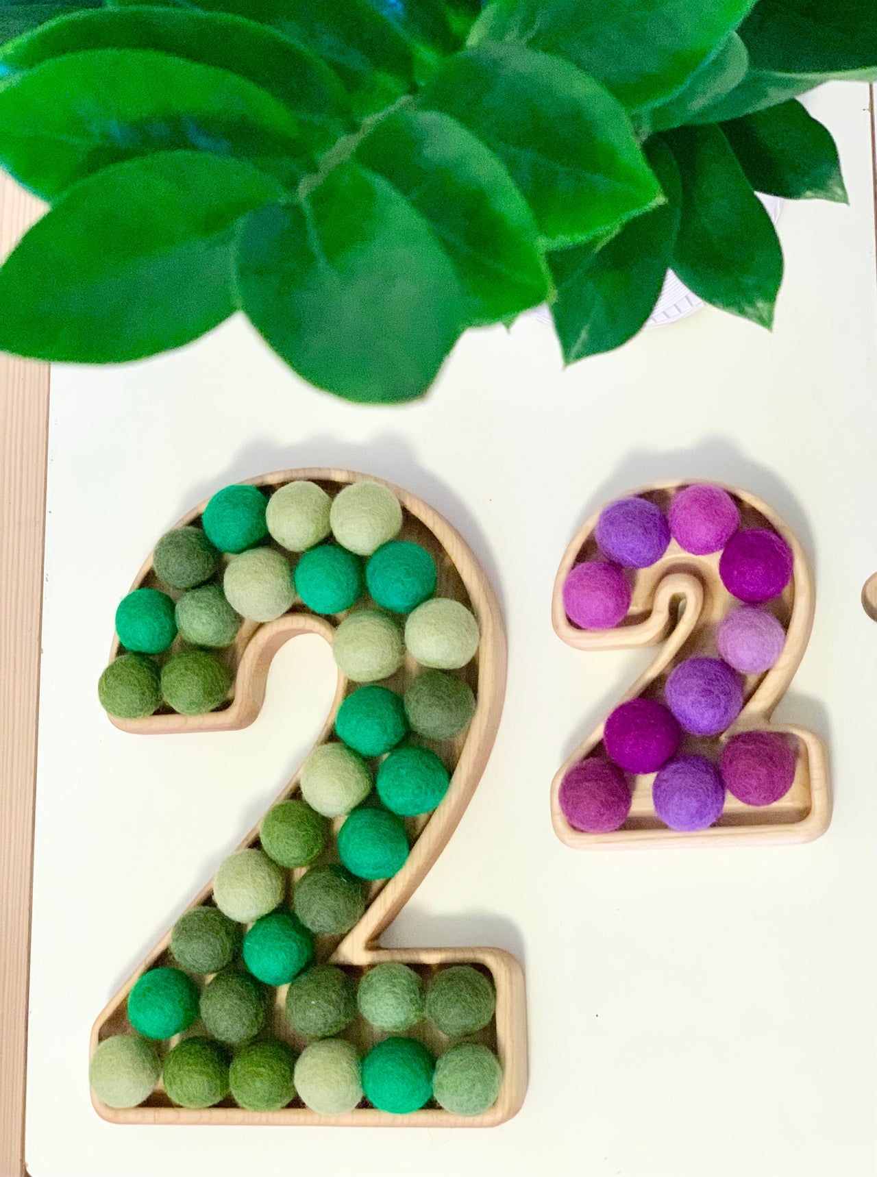 Natural wooden number plate shaped like number 2 filled with green felt balls and smaller number 2 with purple felt balls on white surface