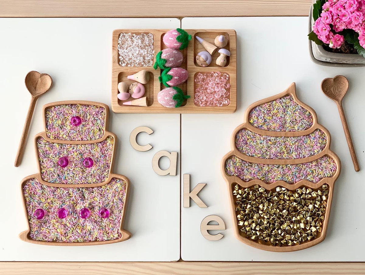 Wooden sensory trays shaped like a layered cake and cupcake filled with colorful sensory materials on white surface