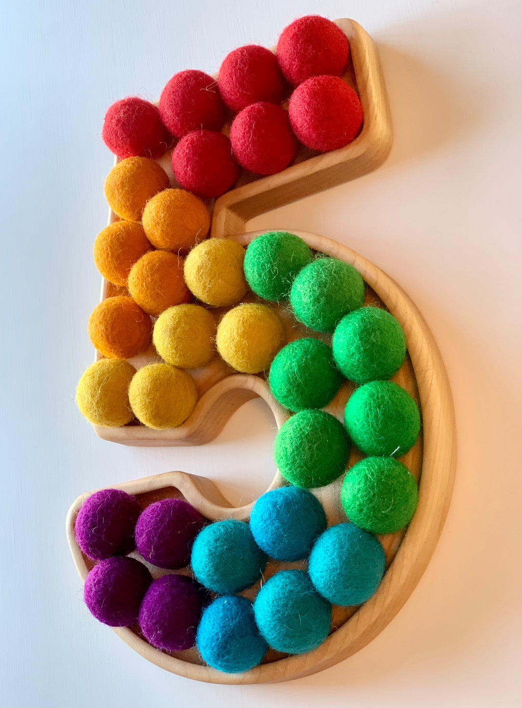 Wooden number-shaped sensory tray filled with colorful felt balls arranged by rainbow colors on white surface