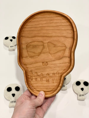 Hand-carved wooden skull-shaped sensory tray with engraved facial features held over white surface with plush skull toys