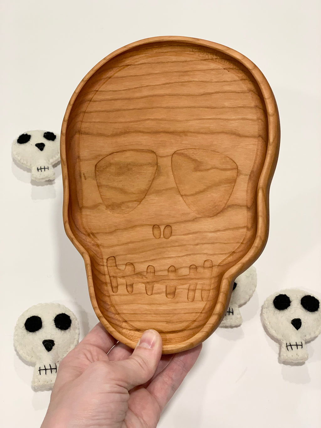 Hand-carved wooden skull-shaped sensory tray with engraved facial features held over white surface with plush skull toys
