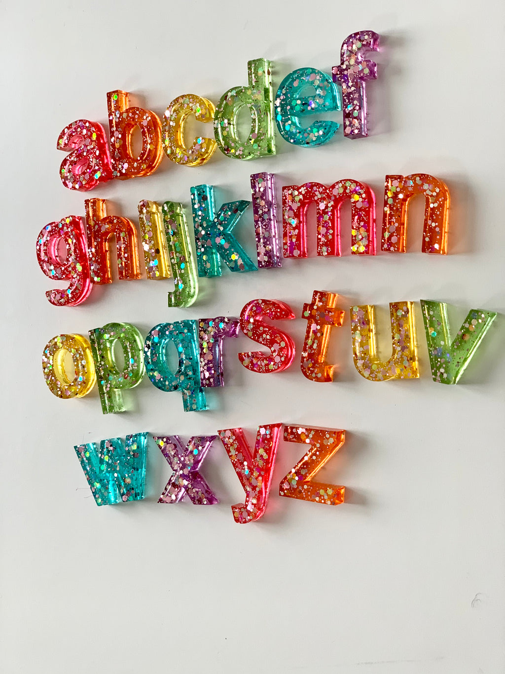 Rainbow glitter resin lowercase alphabet letters arranged in rows on white surface