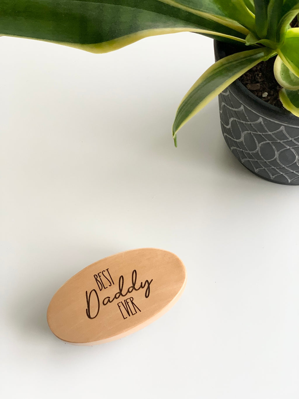 Oval wooden beard brush engraved with "Best Daddy Ever" text on white surface next to green potted plant