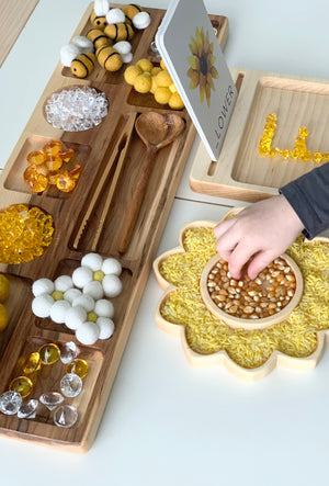 Wooden compartment tray with sensory play items including felt balls, gems, wooden tools, and sunflower-shaped sensory tray with yellow rice and kernels
