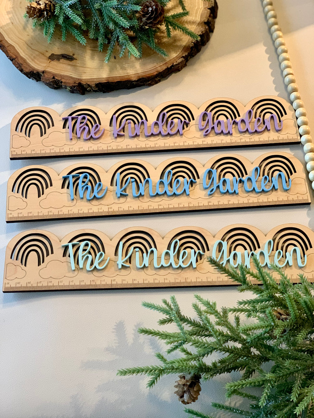 Three personalized wooden rainbow rulers with "The Kinder Garden" in purple, blue, and light blue on natural wood background