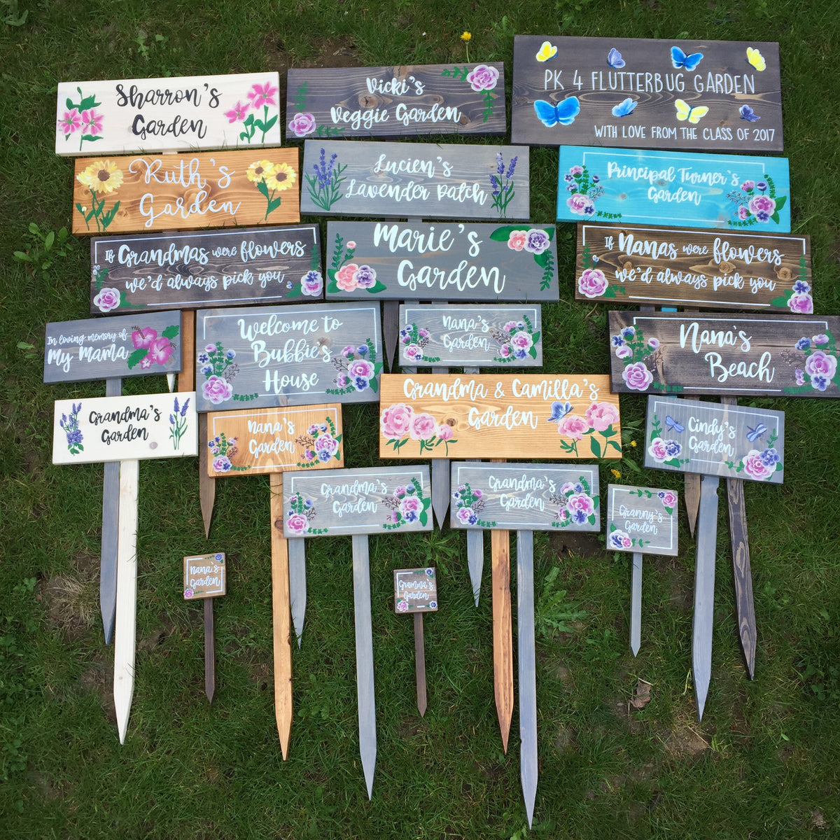 Collection of personalized wooden garden signs with white cursive letters and colorful floral illustrations on grass background