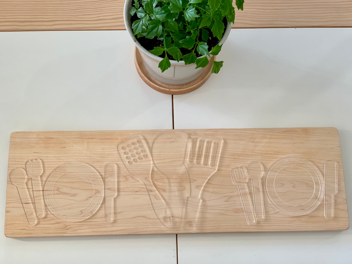 Maple plywood flat dish organizer with engraved spoon, fork, knife, spatula, and plate shapes on white table with plant above
