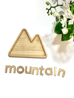 Natural wooden mountain-shaped sensory tray with wooden letters spelling mountain on white background next to flowers