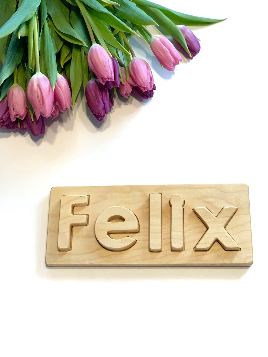 Personalized hardwood name puzzle with lowercase letters spelling FELIX on natural wood board with purple tulips nearby
