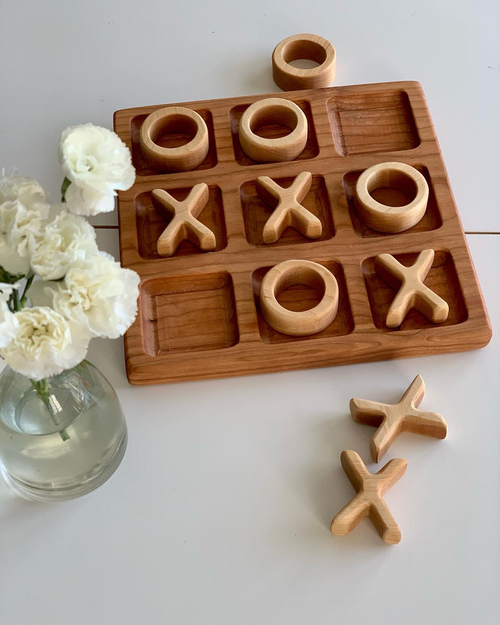 Cherry wood 3x3 tic tac toe board with carved X and O pieces arranged in grid on white surface next to white flowers in vase