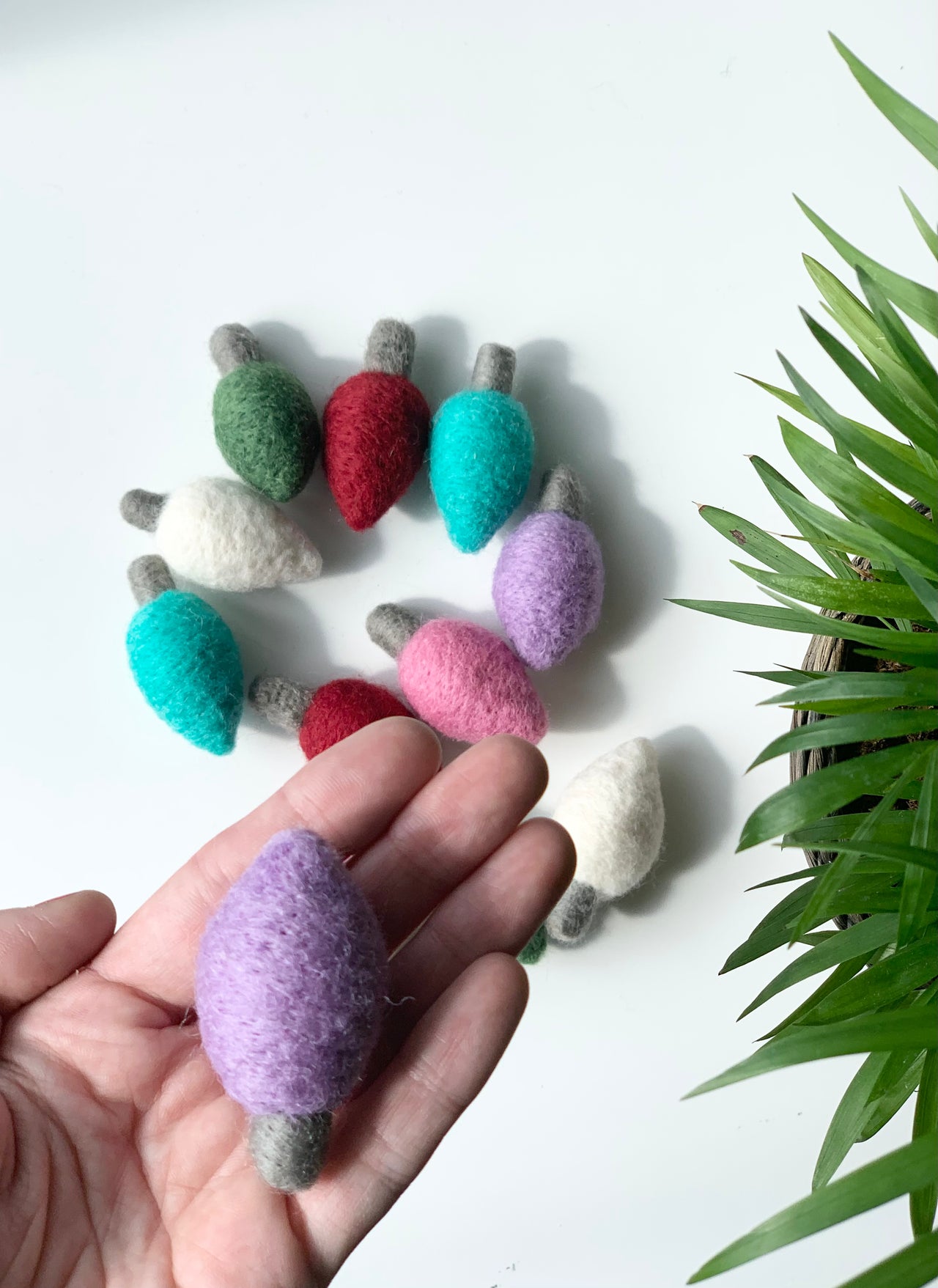 Hand holding a single lavender felt light bulb with assorted colorful felt bulbs arranged on white surface next to green plant