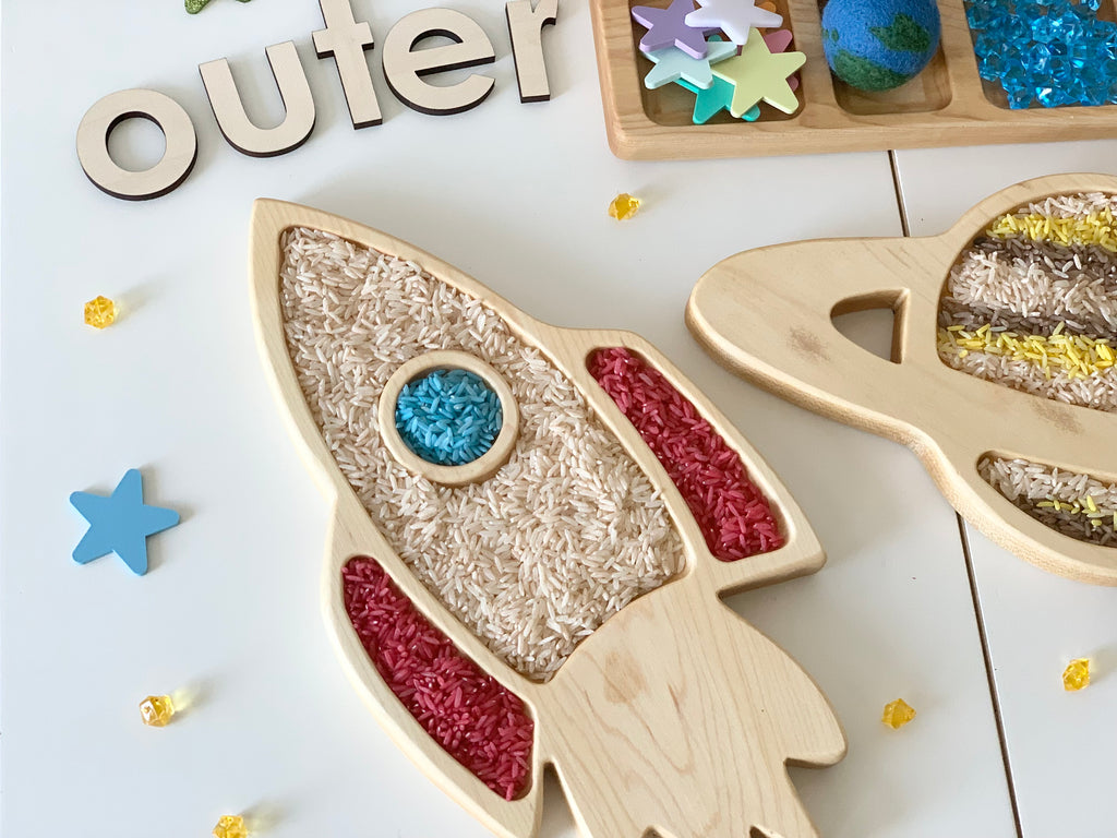 Natural wooden rocket ship sensory tray filled with colored rice on white surface with star toys and wooden letters