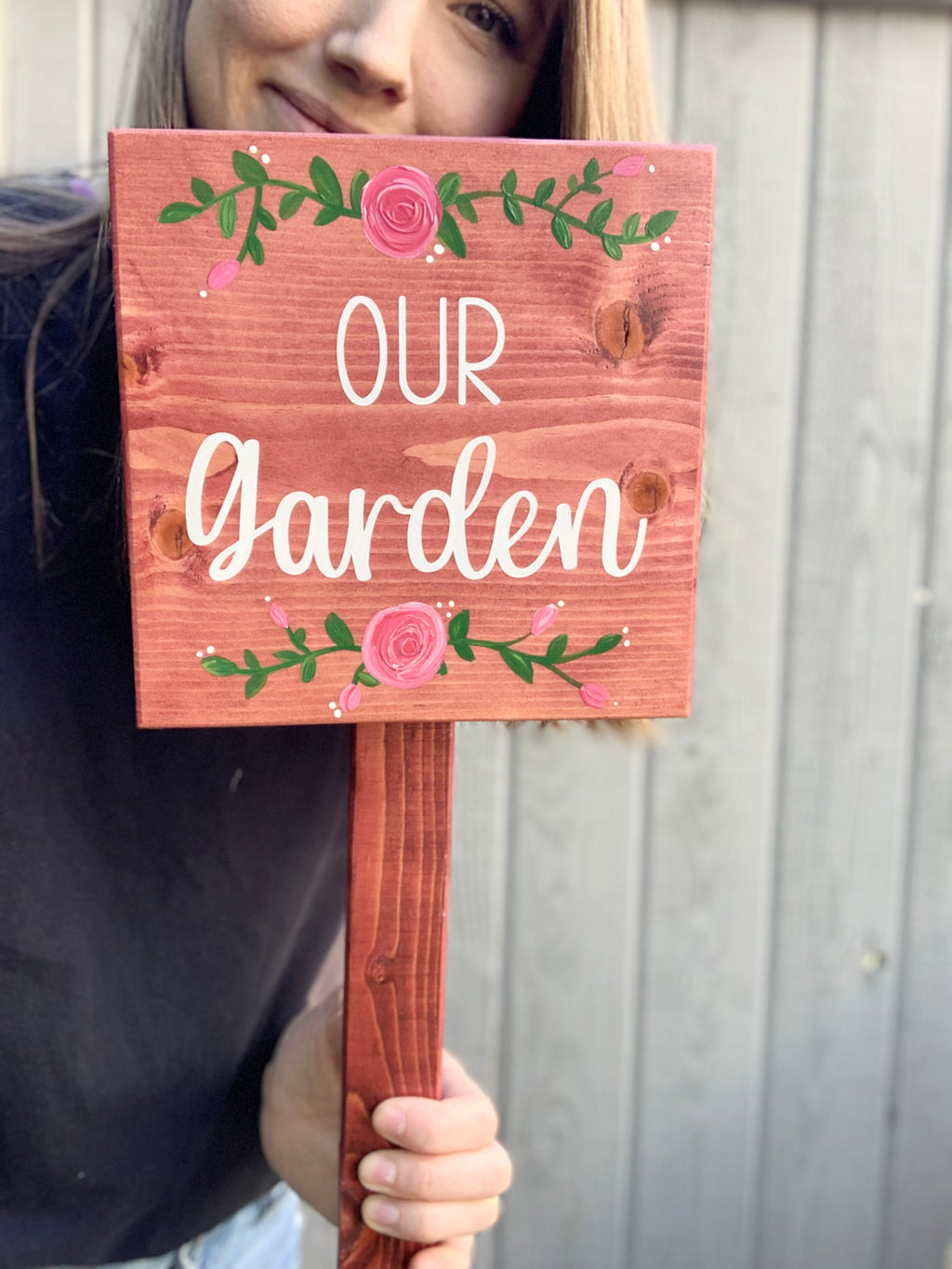 Small square wooden garden sign with white "Our garden" text and pink floral designs on natural wood background held by person