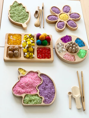 Set of wooden sensory trays shaped like unicorn, flower, leaf, and divided tray filled with colorful textured materials on white surface