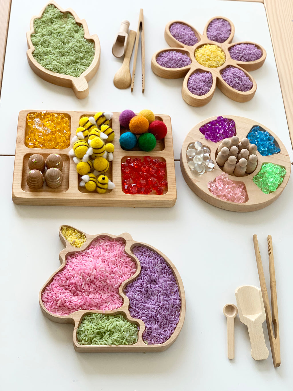 Set of wooden sensory trays shaped like unicorn, flower, leaf, and divided tray filled with colorful textured materials on white surface