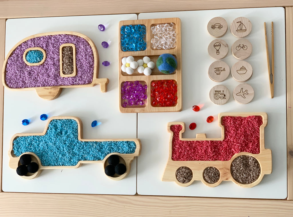 Wooden train and truck sensory trays with colorful textured fill and wooden tokens engraved with vehicle illustrations on white surface