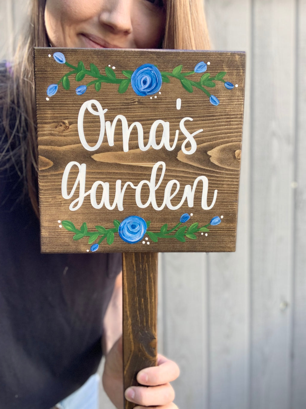 Small square wooden garden sign with white cursive letters and blue floral accents on stained wood background