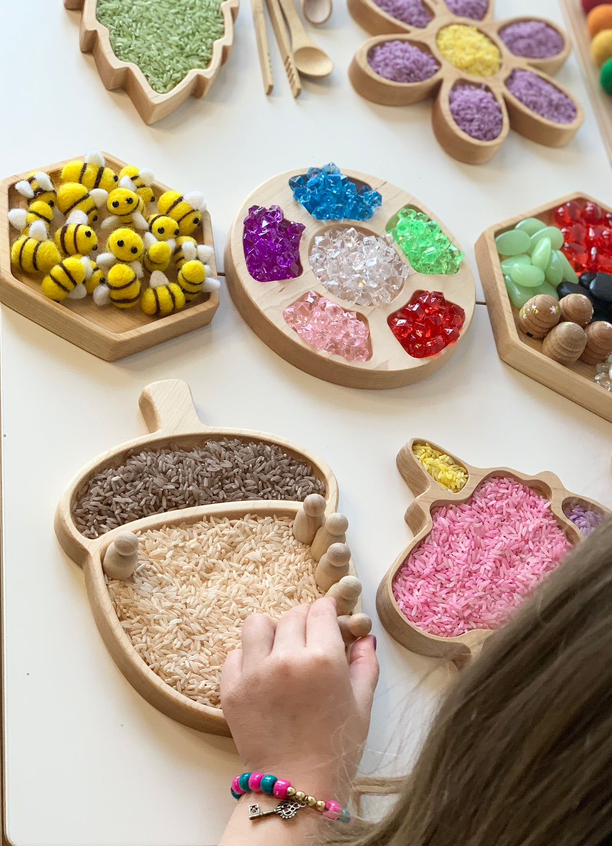 Wooden acorn-shaped sensory tray filled with colored rice next to flower and bee-shaped trays with various sensory materials