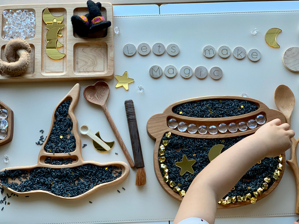 Wooden witch hat and cauldron sensory trays filled with black rice and gold star decorations on white surface with child’s hand
