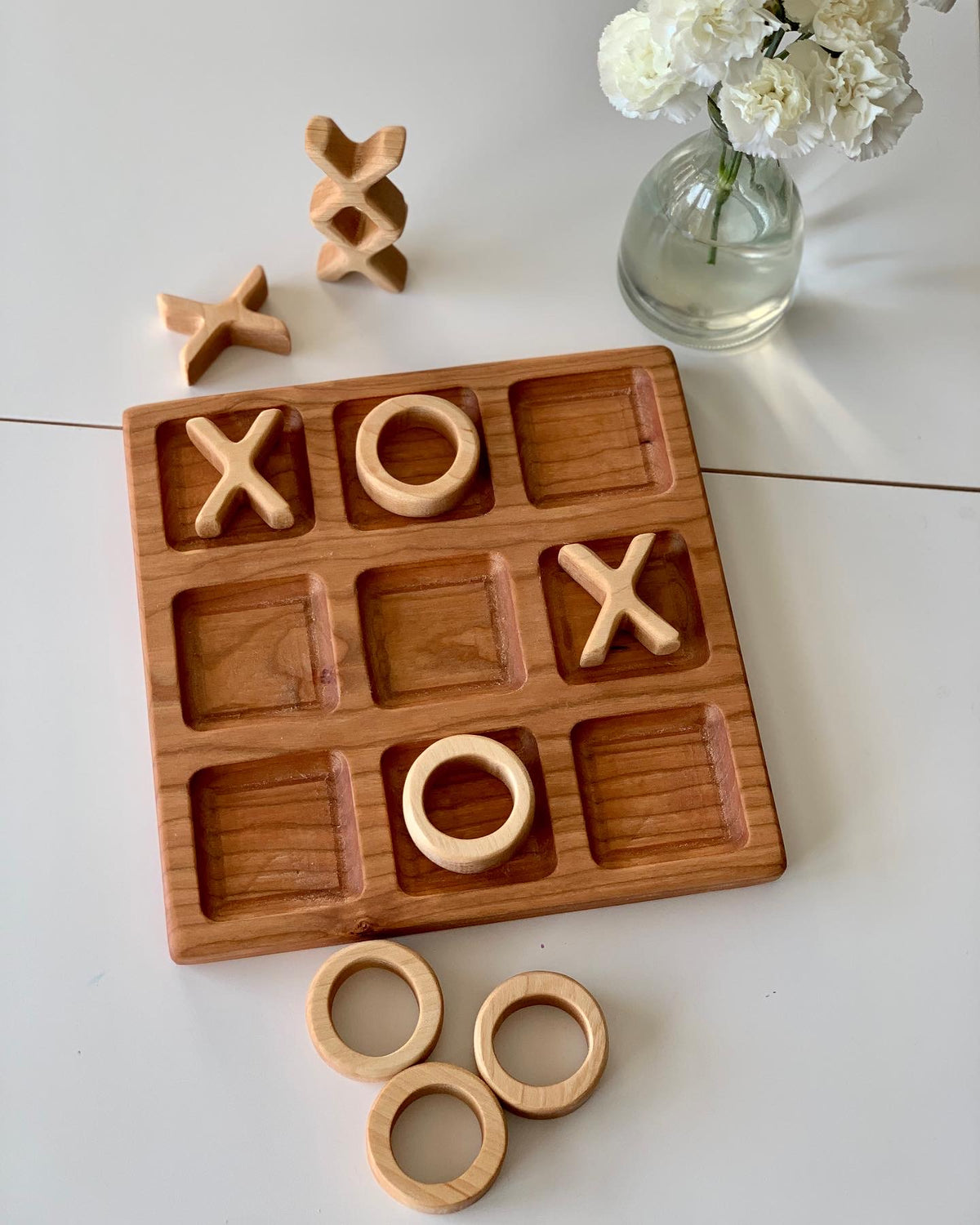 Wooden 3x3 tic tac toe board in cherry wood with carved X and O pieces arranged and stacked on white surface near white flowers