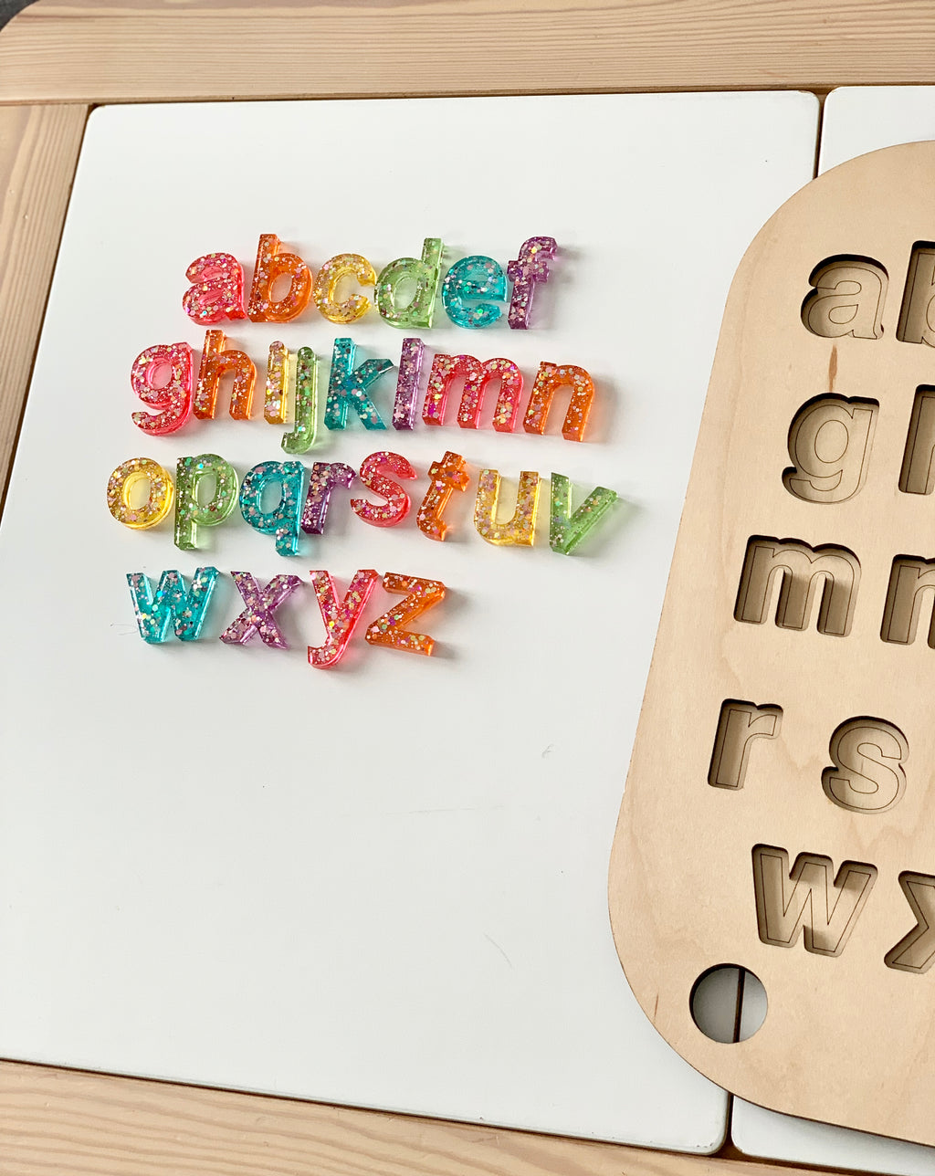Rainbow glitter resin lowercase alphabet letters arranged next to wooden letter tray on white surface