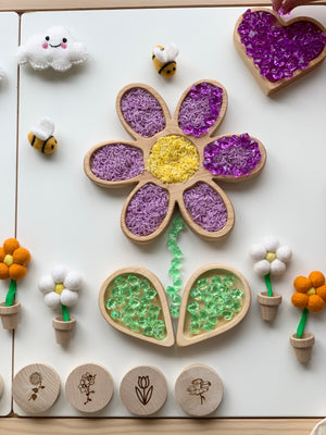 Wooden flower-shaped sensory tray with purple rice, yellow center, green beads, small plush bees and felt flowers on white surface