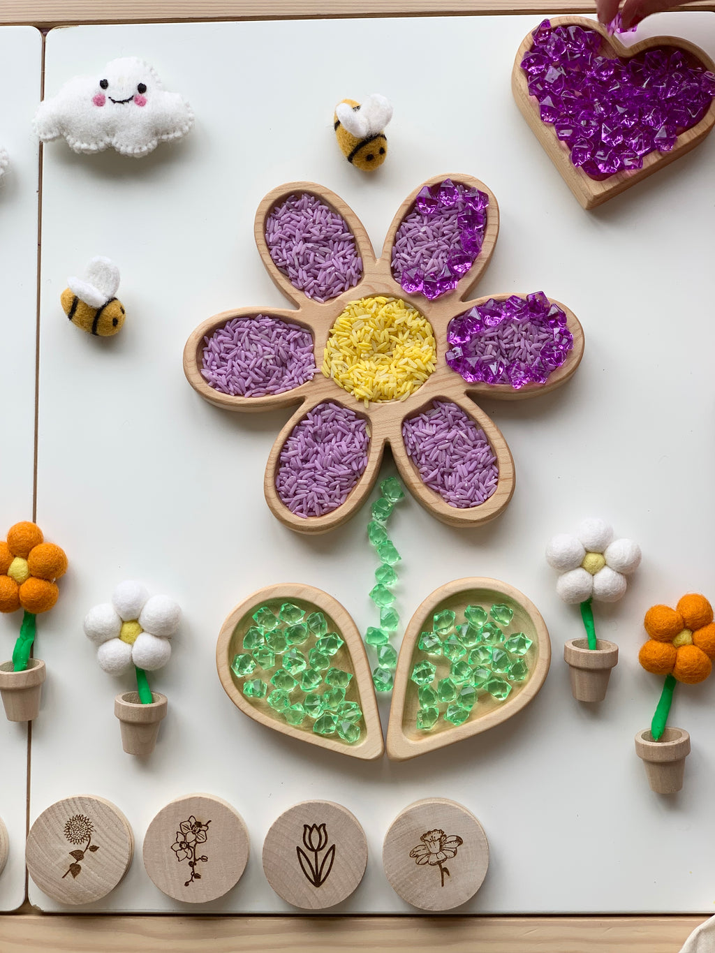 Wooden flower-shaped sensory tray with purple rice, yellow center, green beads, small plush bees and felt flowers on white surface