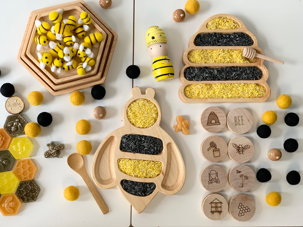 Wooden bee-shaped sensory tray with yellow and black rice, small wooden bee figure, hexagon tray with felt bees, and wooden tokens on white surface