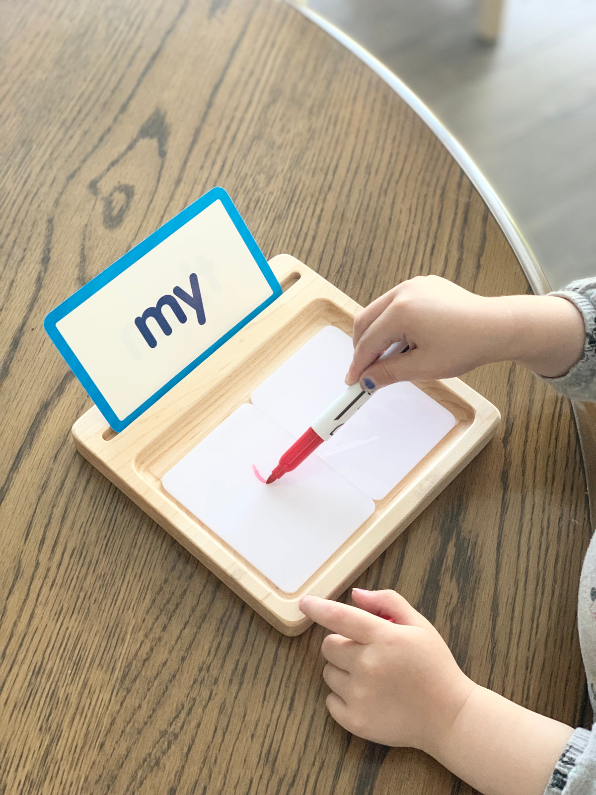 Child using red dry erase marker on white flash card in wooden sensory writing tray with word card my