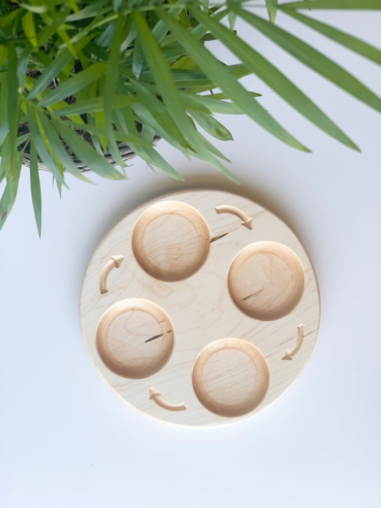 Round natural wood sensory tray with four circular compartments and curved arrows on white surface near green leaves