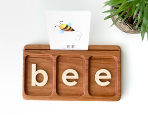 Wooden three-section CVC tray with wooden letters spelling BEE and illustrated flash card of a bee above tray on white surface