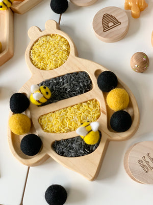 Bee-shaped wooden sensory tray filled with yellow and black rice with felt bee and pom-pom balls nearby