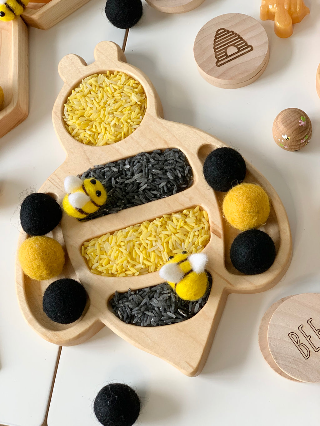 Bee-shaped wooden sensory tray filled with yellow and black rice with felt bee and pom-pom balls nearby
