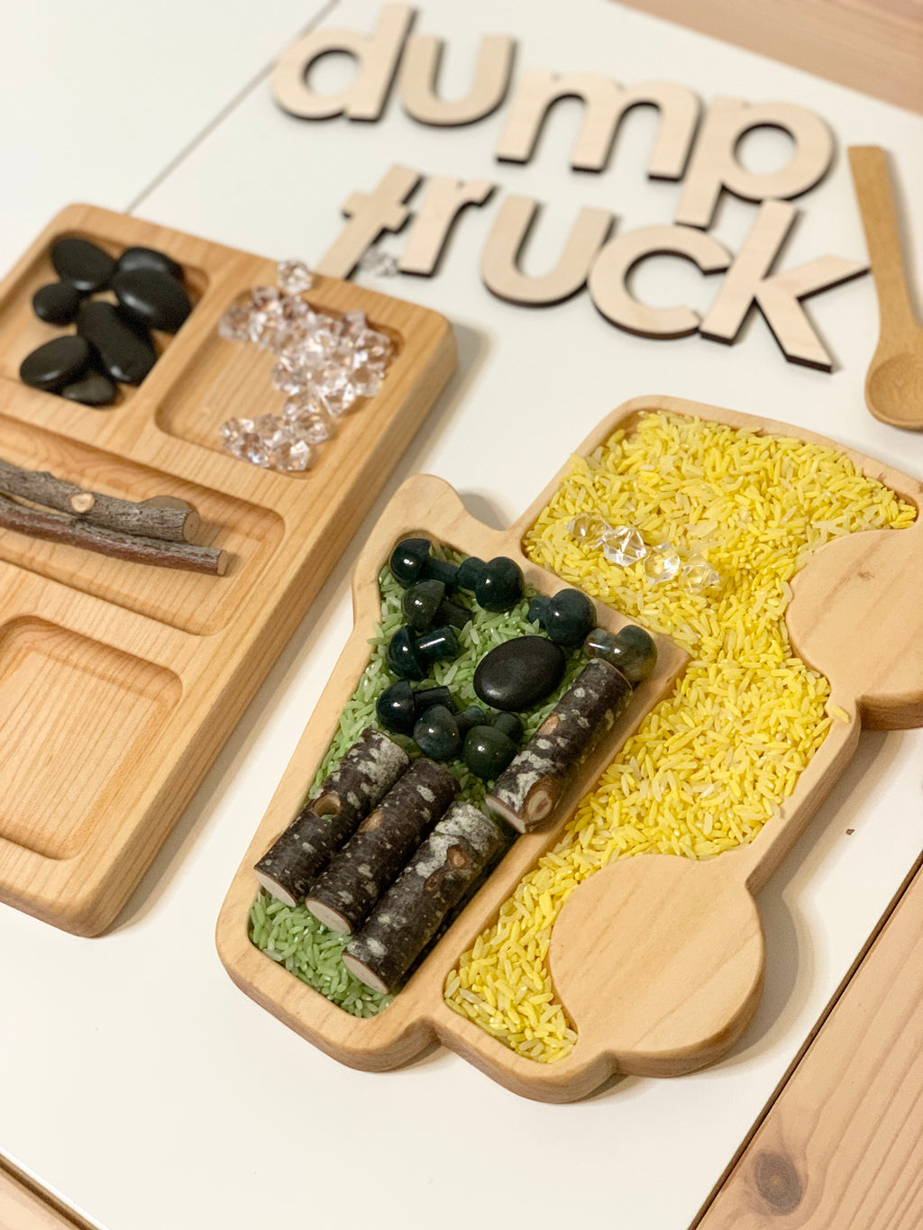 Wooden dump truck sensory tray filled with yellow rice, green rice, small rocks, and wooden sticks on white surface