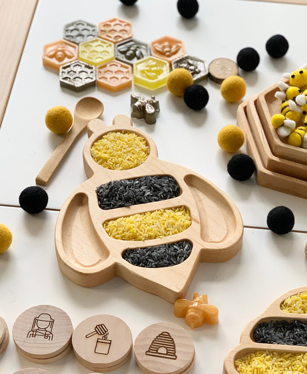 Bee-shaped natural wooden sensory tray filled with yellow and black rice on white surface with small toys