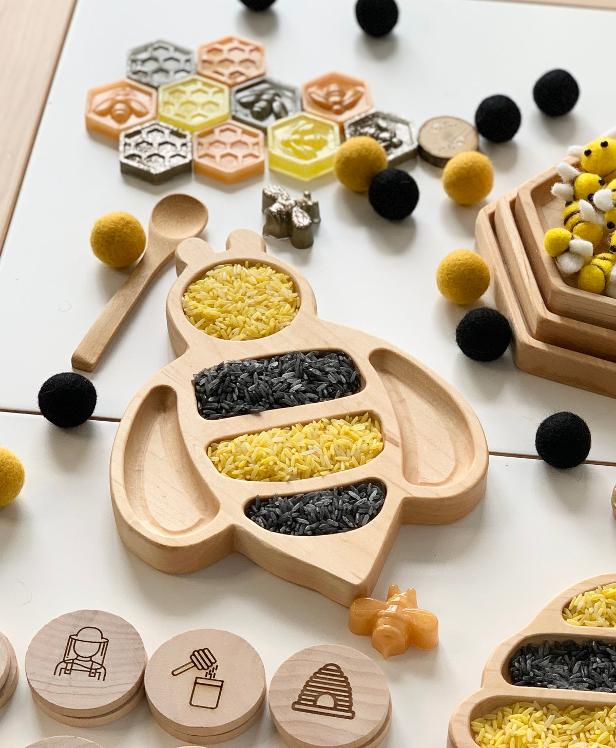 Bee-shaped natural wooden sensory tray filled with yellow and black rice on white surface with small toys