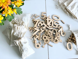 Assorted maple wood numbers and math symbols scattered on white table next to natural drawstring storage bags and yellow flowers