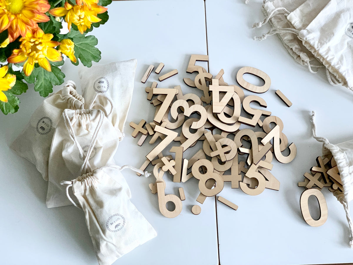 Assorted maple wood numbers and math symbols scattered on white table next to natural drawstring storage bags and yellow flowers