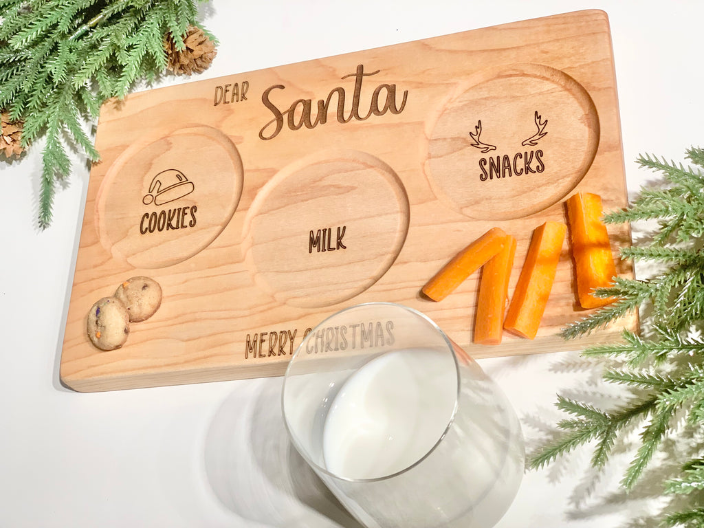 Natural wooden Santa plate with engraved sections for cookies, milk, and reindeer snacks on white background with greenery