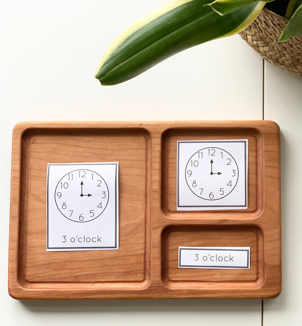 Hardwood three-part card tray with Montessori clock cards showing 3 o’clock on a white surface with plant leaves