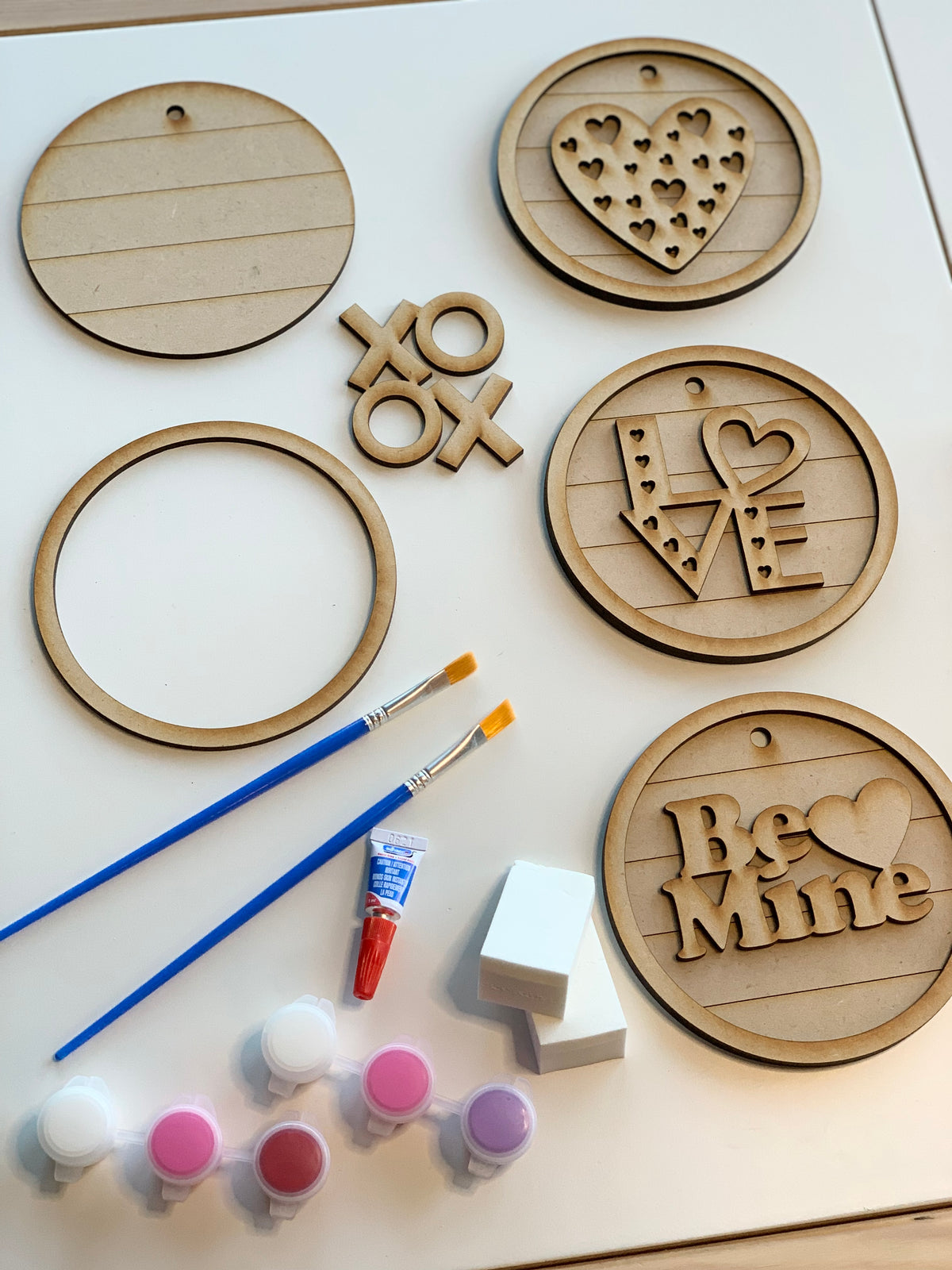 Unpainted wooden circular signs with love-themed designs, paintbrushes, paint pots, and sponges on white surface