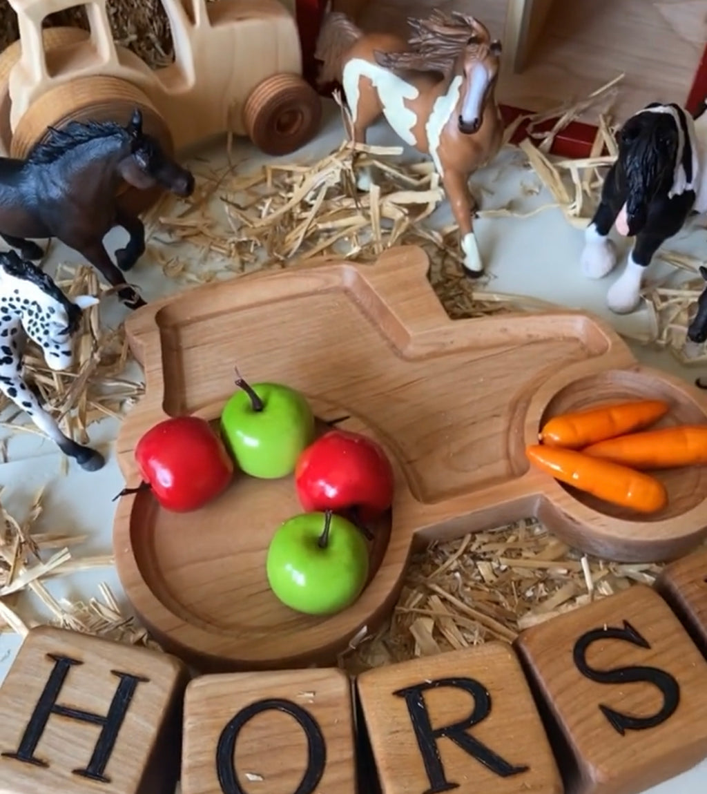 Natural wooden tractor-shaped sensory tray with toy apples and carrots surrounded by toy horses and wooden alphabet blocks