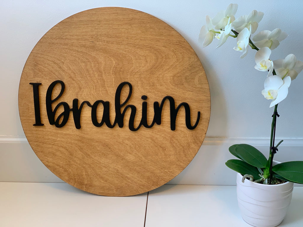 3D round wooden name sign with black cursive letters on natural wood grain background next to white orchid plant